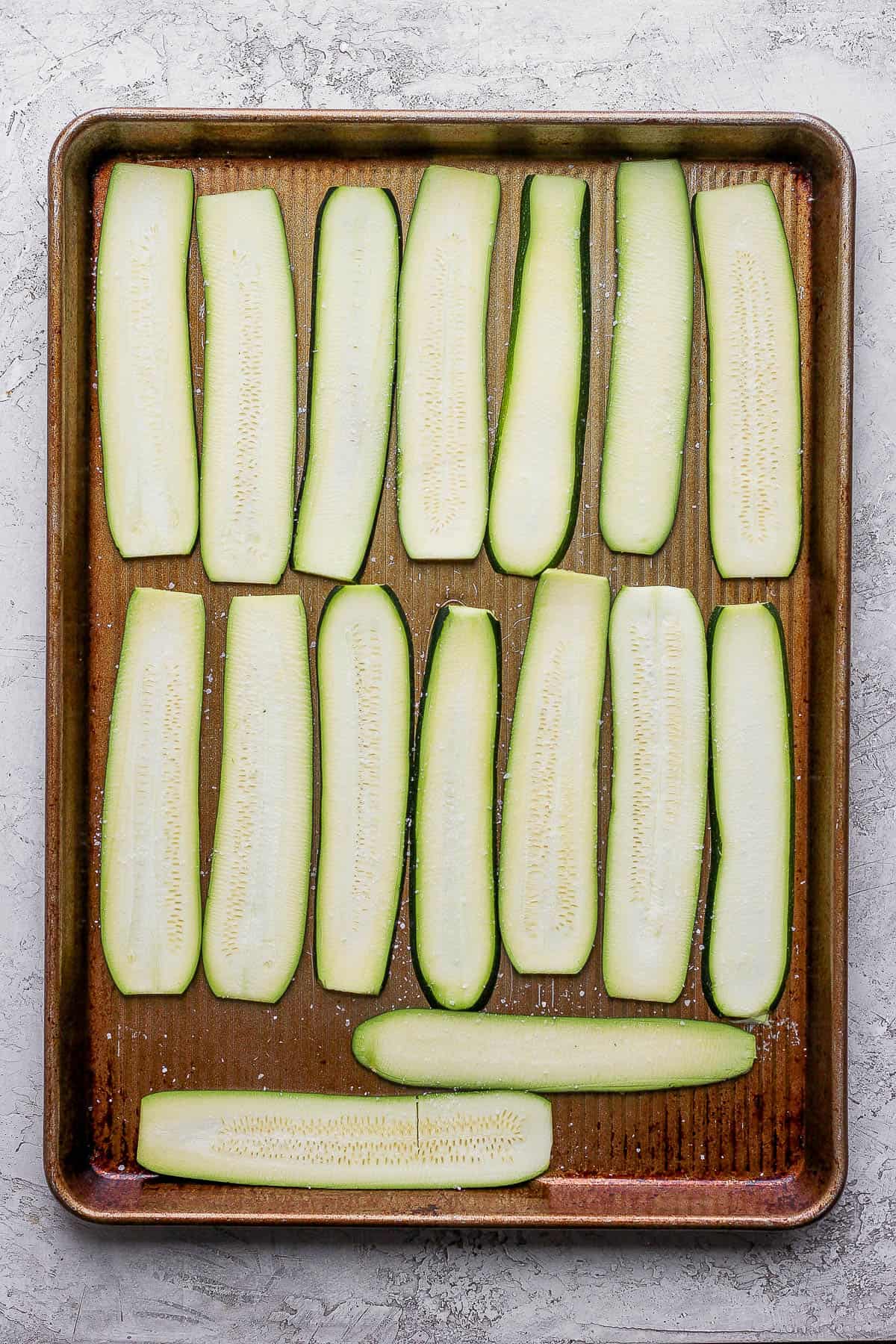 Two rows of horizontally sliced zucchinis laid out evenly on a baking sheet and sprinkled with salt. 