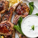 A close up of a plate of lamb chops with a bowl of lamb sauce, mint leaves and lemon slices.