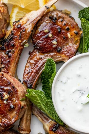 A close up of a plate of lamb chops with a bowl of lamb sauce, mint leaves and lemon slices.