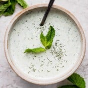 A bowl of homemade tzatziki sauce with a spoon sticking out and some fresh mint leaves on top.