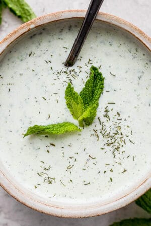 A bowl of homemade tzatziki sauce with a spoon sticking out and some fresh mint leaves on top.