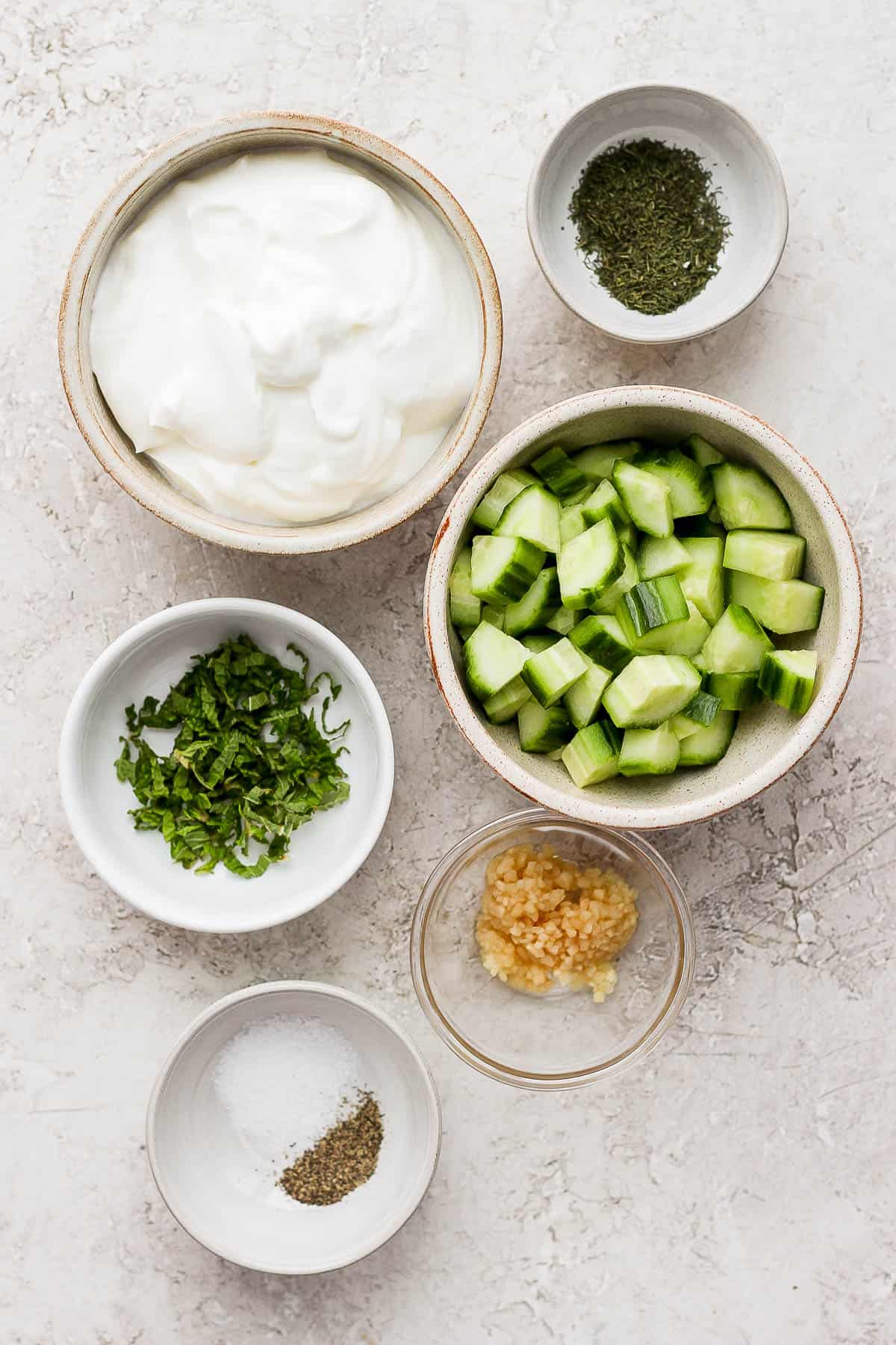 Six small bowls, each holding prepped and measured ingredients for the tzatziki sauce; a bowl of dill, a bowl of Greek yogurt, a bowl of peeled and cut quartered cucumber, a bowl of fresh sliced mint, a bowl of minced garlic, and a bowl of kosher salt and ground black pepper. 
