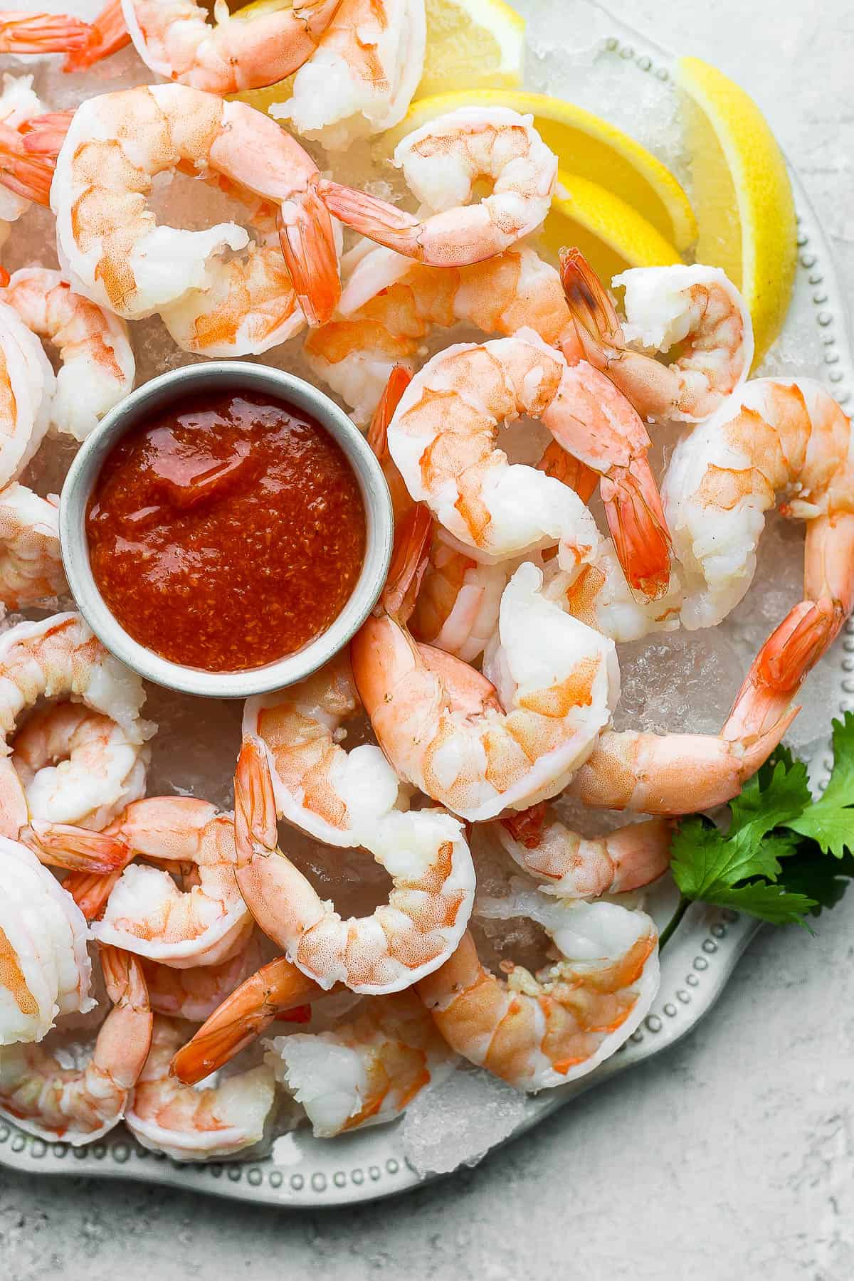 A white platter lined with ice cubes with shrimp spread evenly over the top.  A small bowl of cocktail sauce is nestled between the shrimp on the ice lined platter.