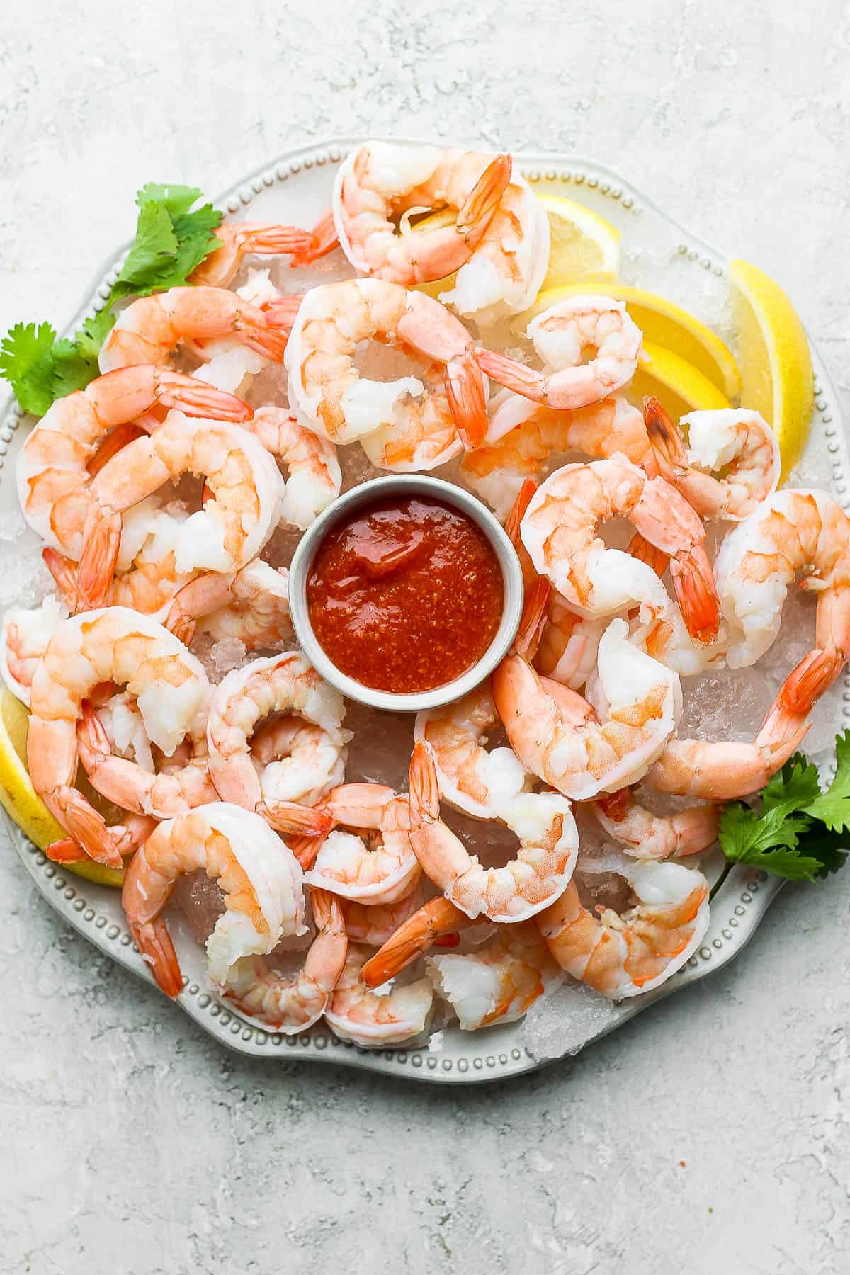 A white serving platter lined with ice cubes with shrimp placed on top with a bowl of cocktail sauce nestled in the middle.  There are lemon wedges on the side of the platter.