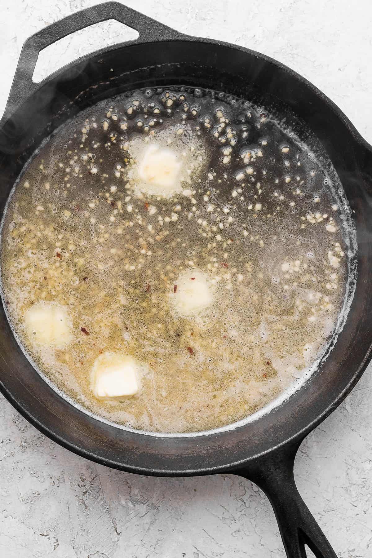 A large cast iron skillet with olive oil, garlic, butter, wine, salt, pepper, and red pepper flakes.