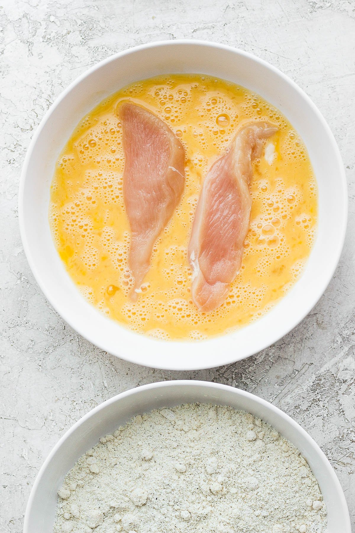 Two white bowls, one with chicken tenders in the egg wash and a bowl of dry almond flour ingredients.