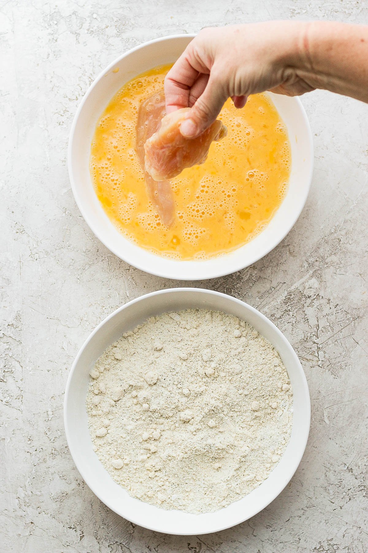 A hand pulling chicken tenders out of the egg wash and another bowl of dry ingredients.