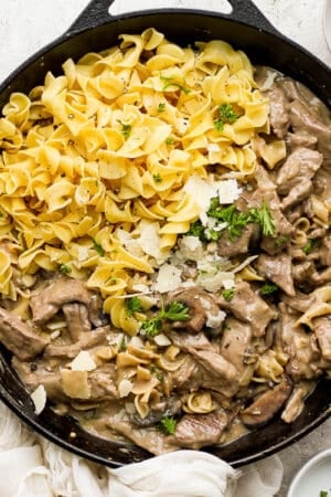 A cast iron skillet filled with beef stroganoff, egg noodles and garnished with parmesan cheese and chopped parsley.
