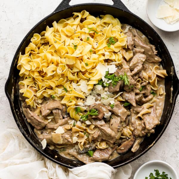 A cast iron skillet filled with beef stroganoff, egg noodles and garnished with parmesan cheese and chopped parsley.