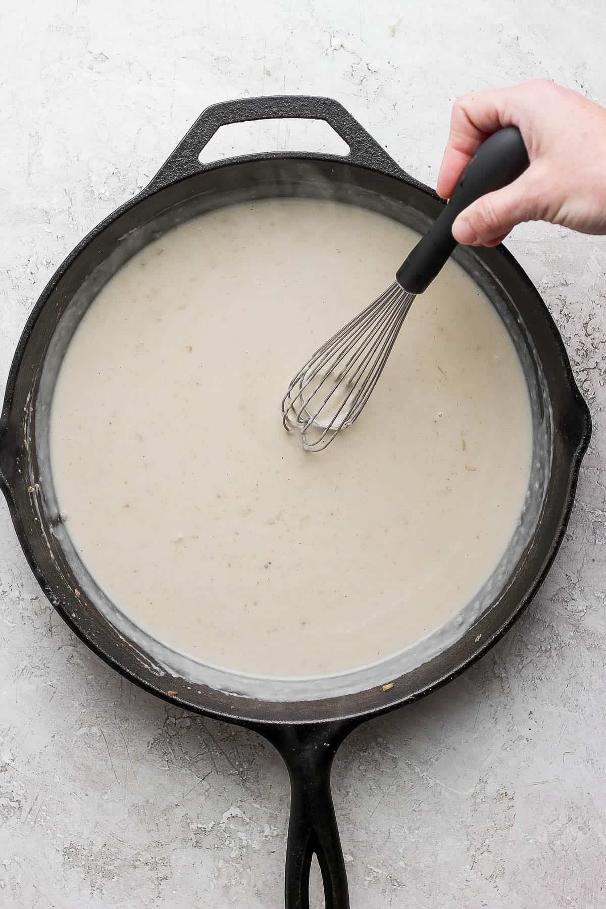 A hand holding a whisk in the cast iron skillet which is filled with the roux .