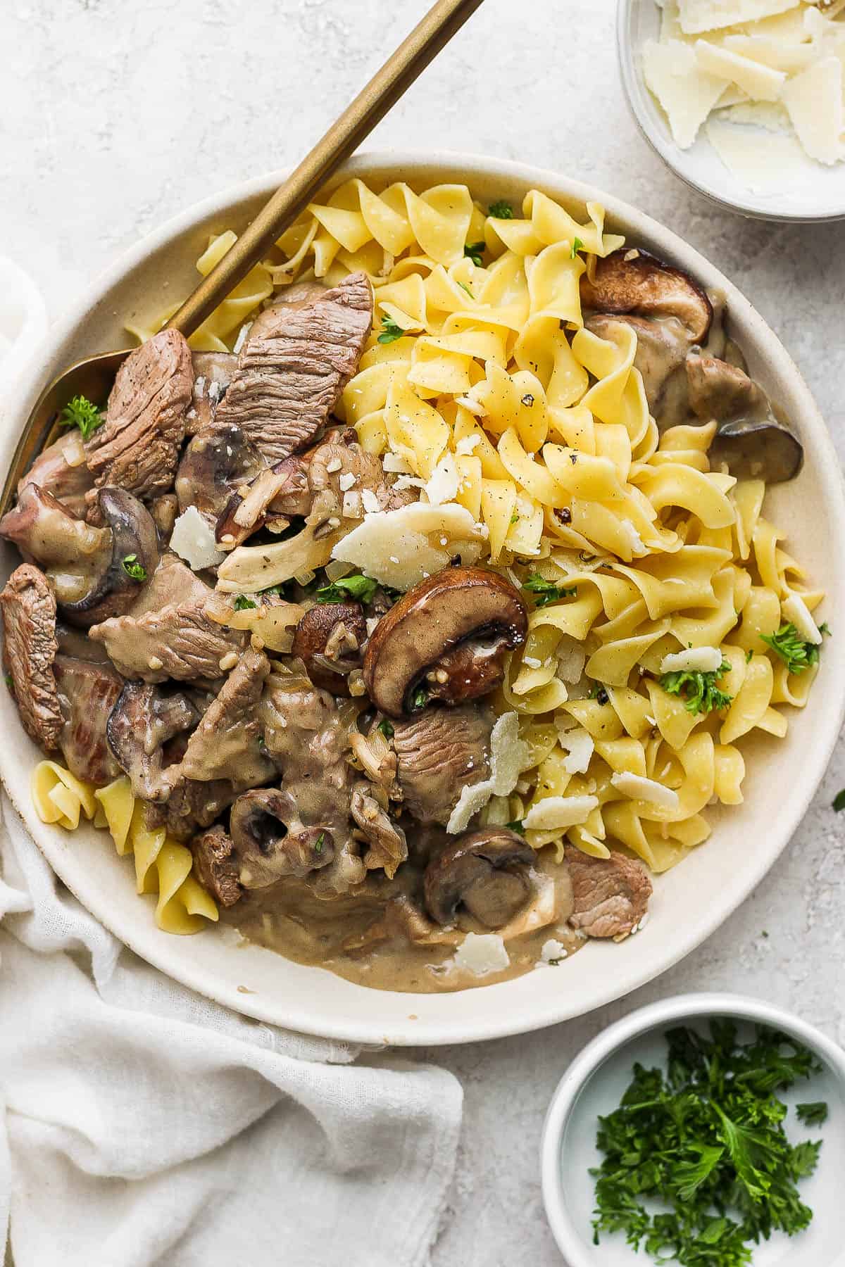 A bowl containing beef stroganoff and cooked egg noodles, all topped with shaved parmesan and freshly chopped parsley.