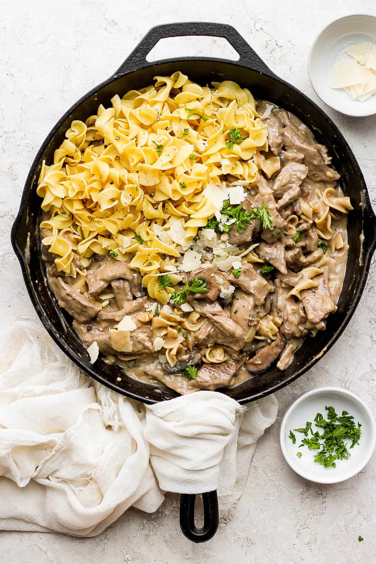 A cast iron skillet with one half filled with cooked egg noddles and the other half filled with cooked beef stroganoff sauce and beef strips, all topped with shaved parmesan and freshly chopped parsley.