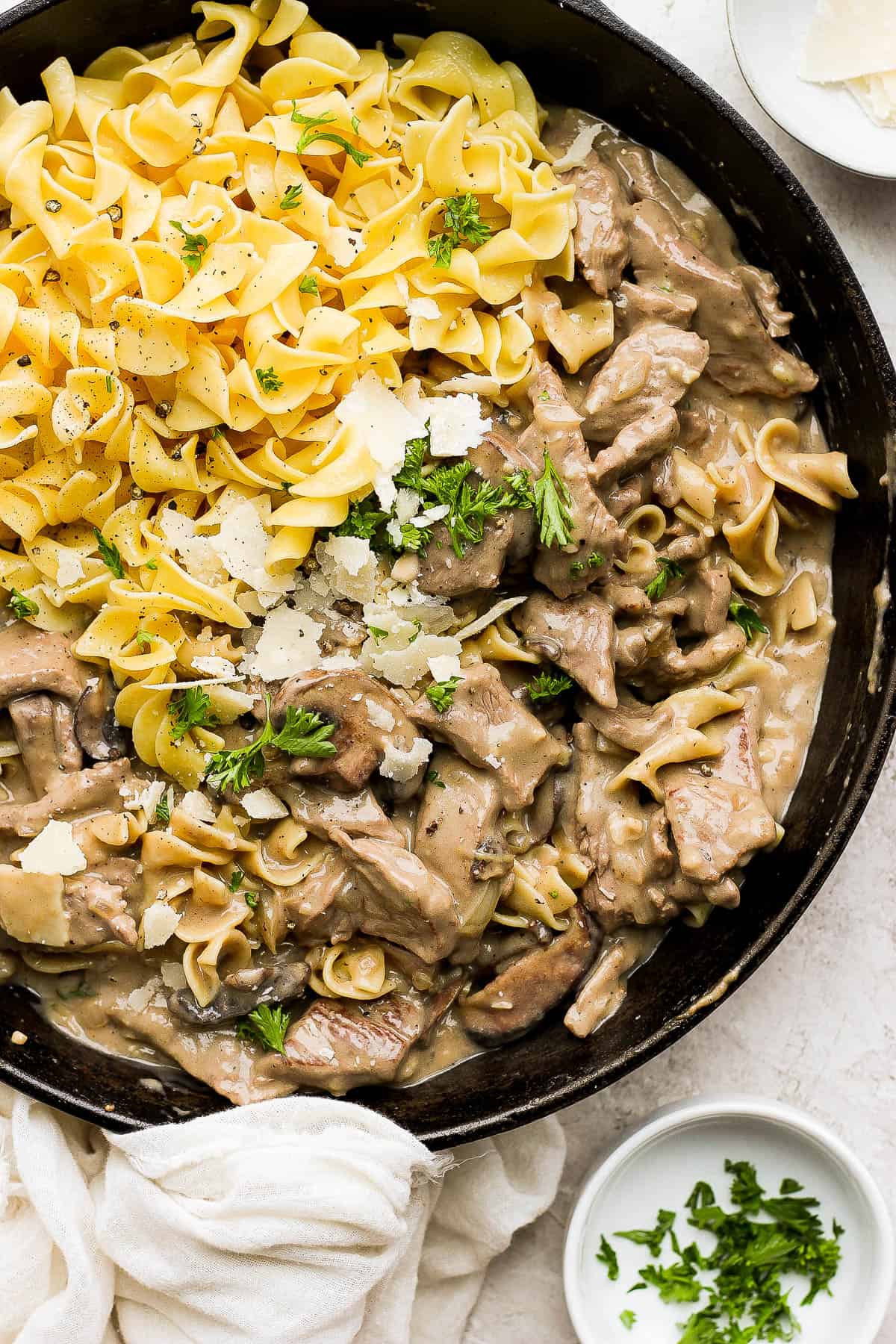 A cast iron skillet with beef stroganoff and cooked egg noodles all topped with shaved parmesan cheese and freshly chopped parsley. 