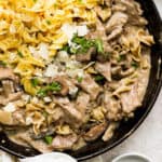 A cast iron skillet filled with beef stroganoff, egg noodles and garnished with parmesan cheese and chopped parsley.