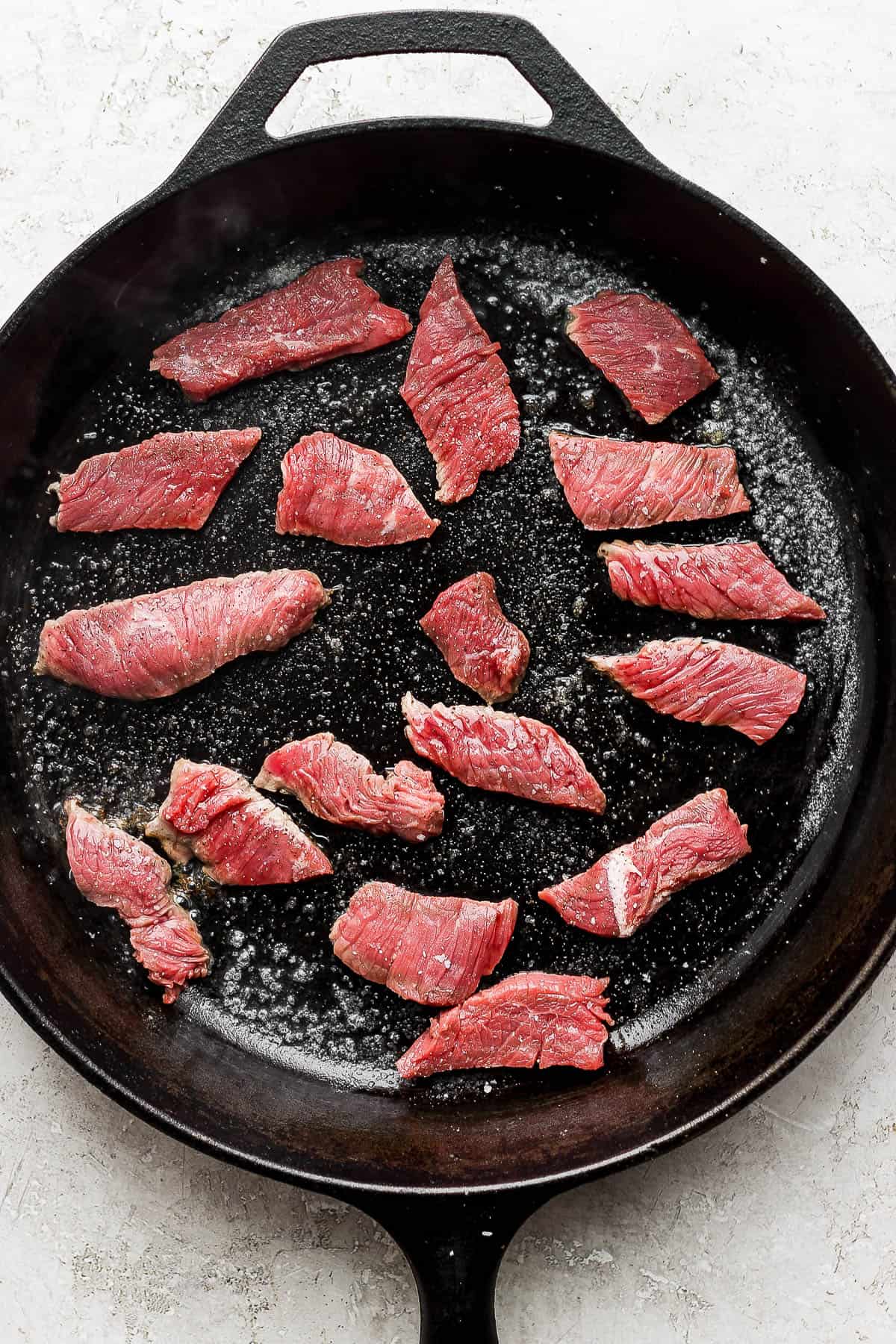 2-inch strips of sirloin steak evenly distributed on a cast iron steak getting seared on each side.