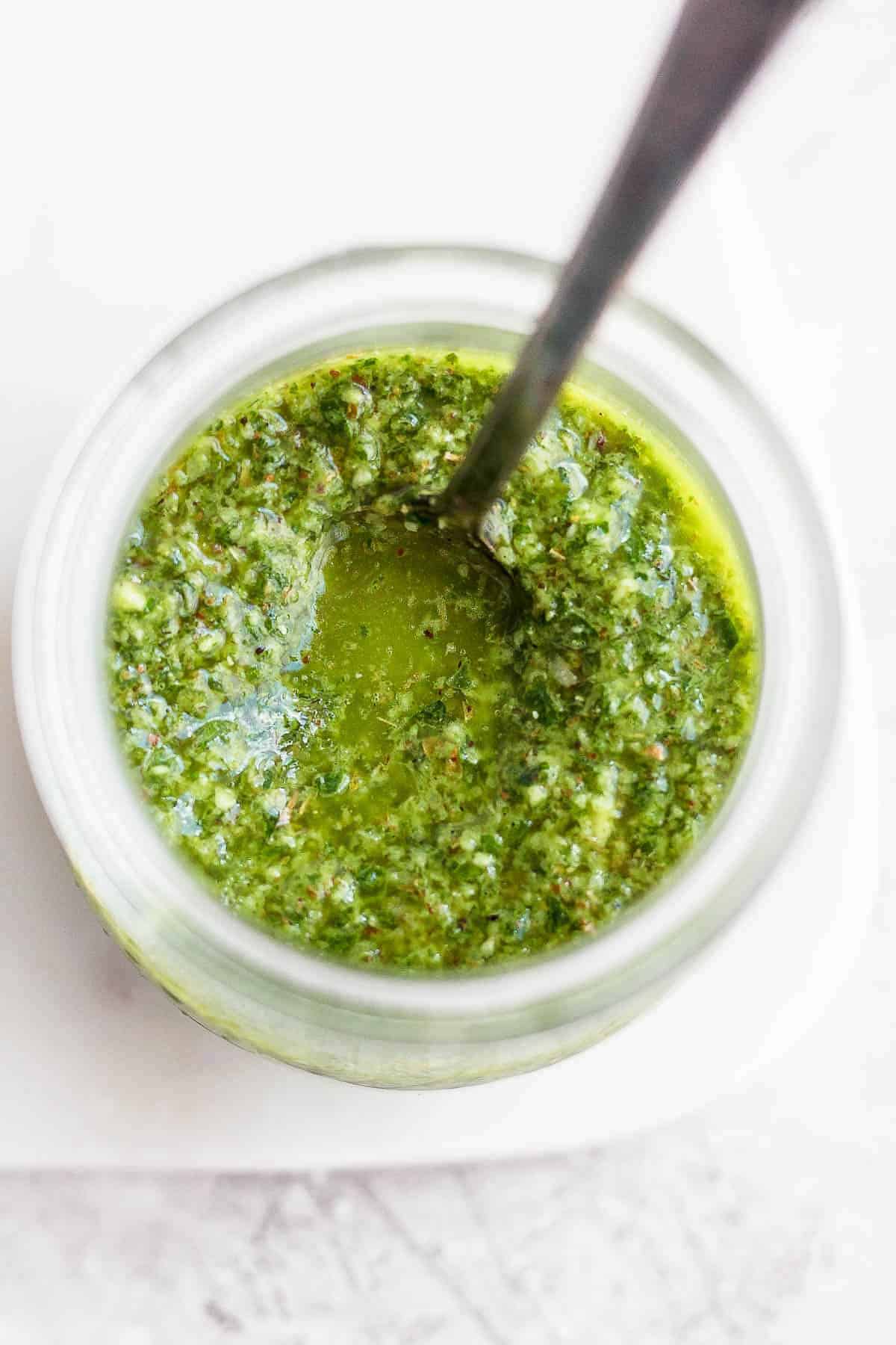 Chimichurri sauce in a glass jar with a spoon inside it.