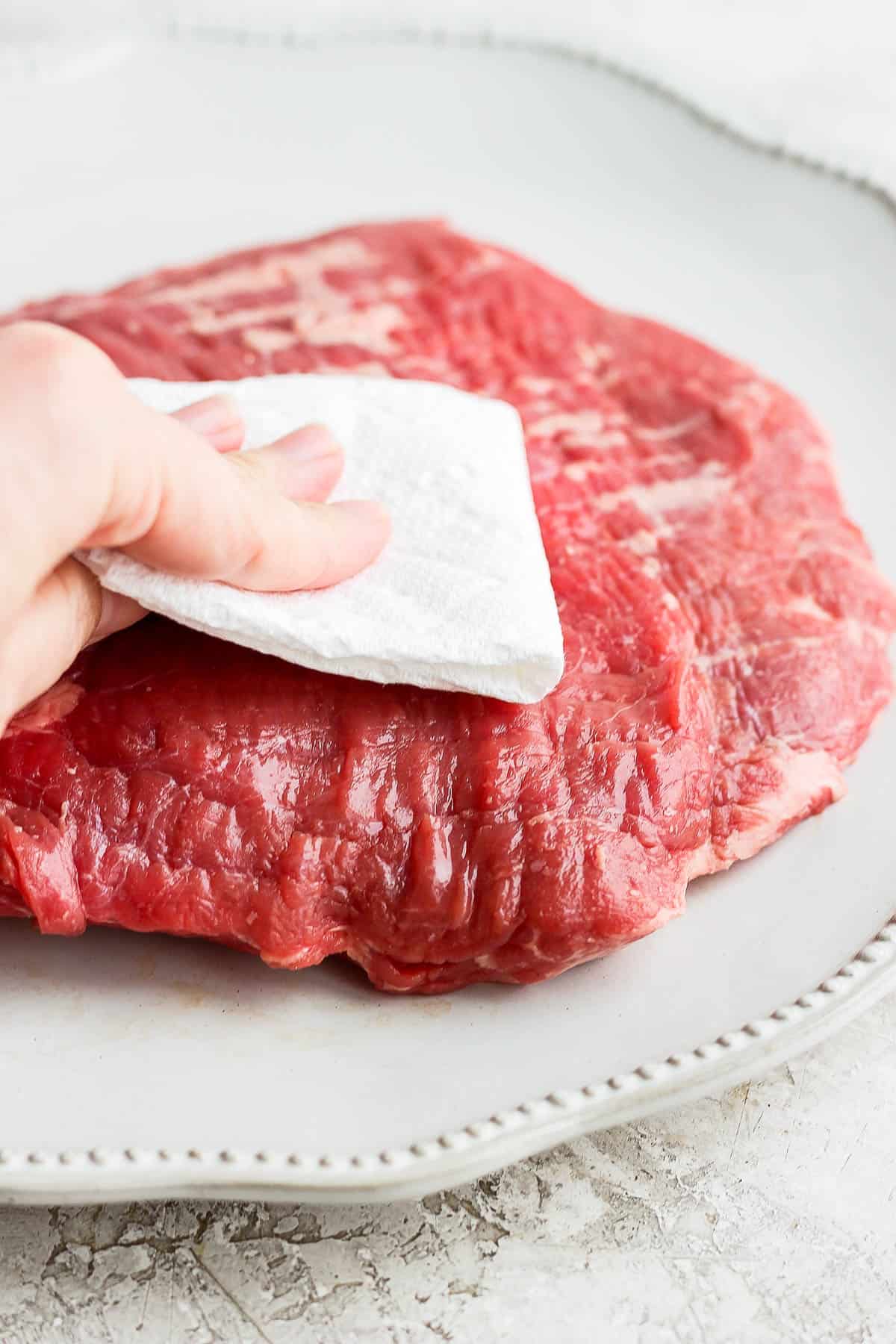 A hand holding a clean paper towel patting a raw piece of flank steak that's sitting on a plate. 