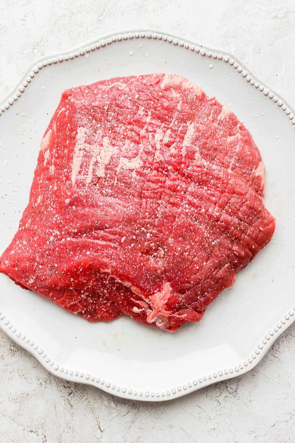 A raw flank steak sprinkled with salt and pepper on a white plate.