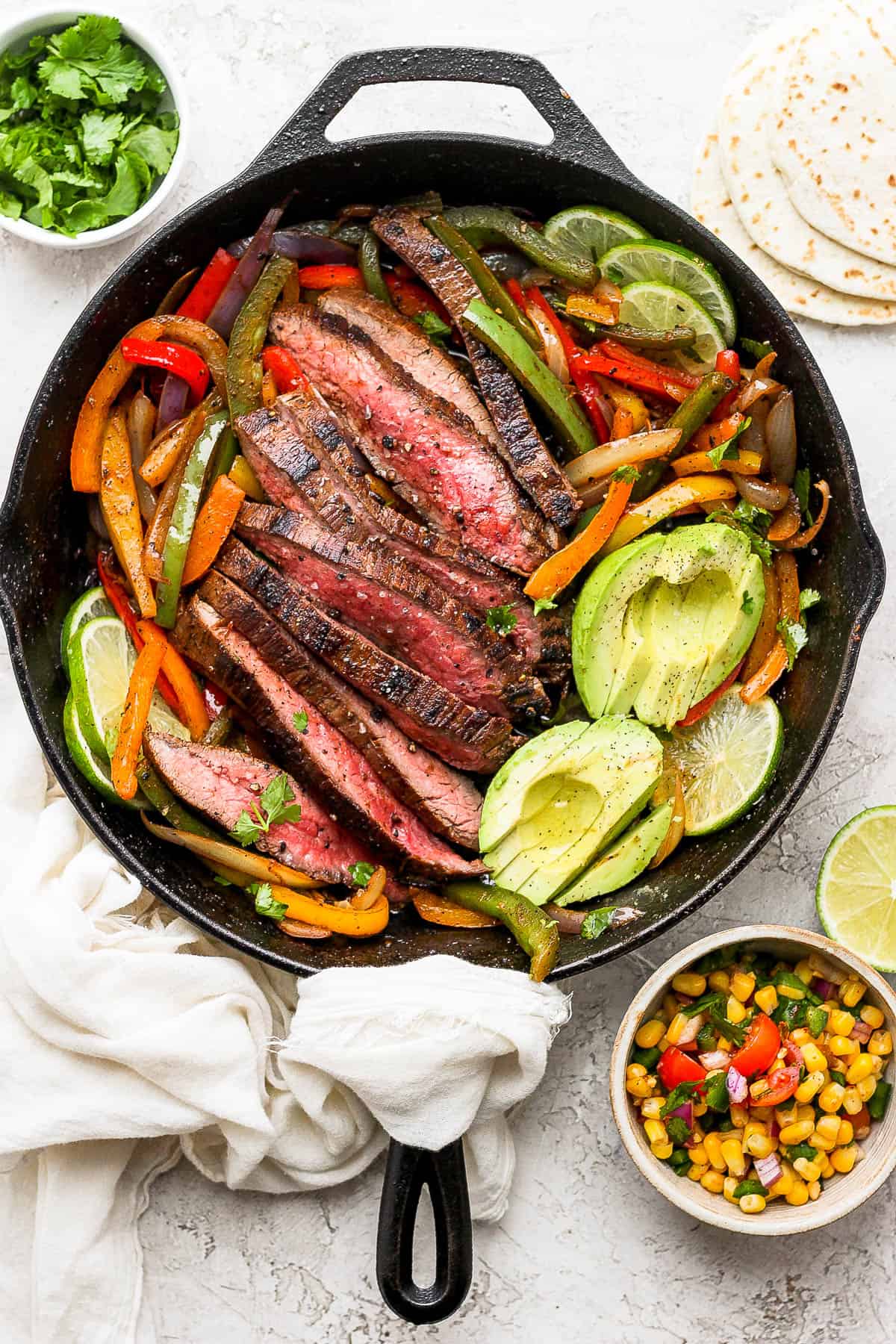 Cooked and sliced flank steak, sautéd bell peppers and onions, slices of lime, and sliced avocado in a cast iron skillet. A bowl of corn salsa is off to the side of the skillet. 