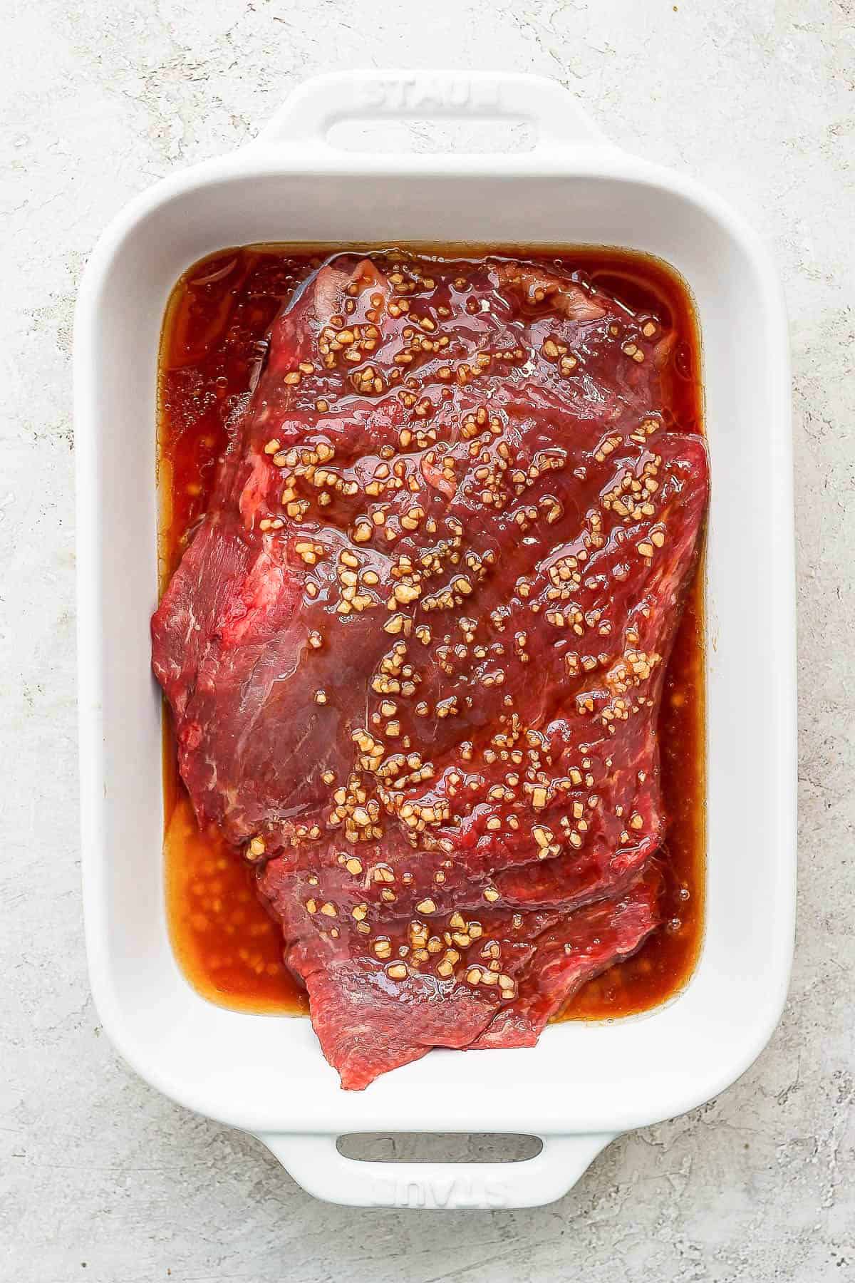 Flank steak covered in marinade in a white casserole dish.