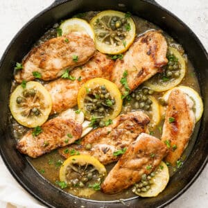 Cast iron skillet filled with chicken piccata with capers and lemon slices.