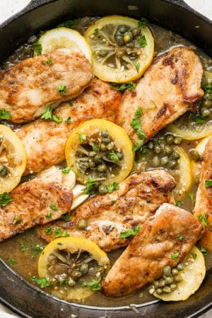 Cast iron skillet filled with chicken piccata with capers and lemon slices.