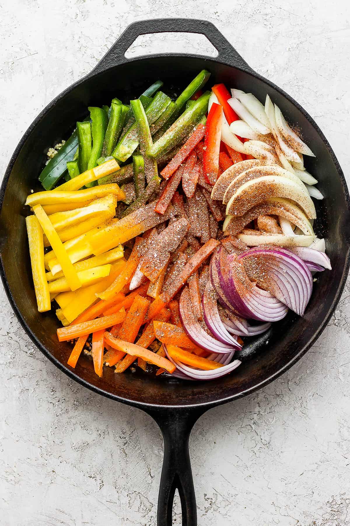 The fajita seasoning sprinkled over sliced bell peppers, onions, and red onion in a cast iron skillet. 
