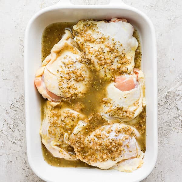 A white baking dish filled with raw bone-in, skin-on chicken thighs covered in a Greek chicken marinade.