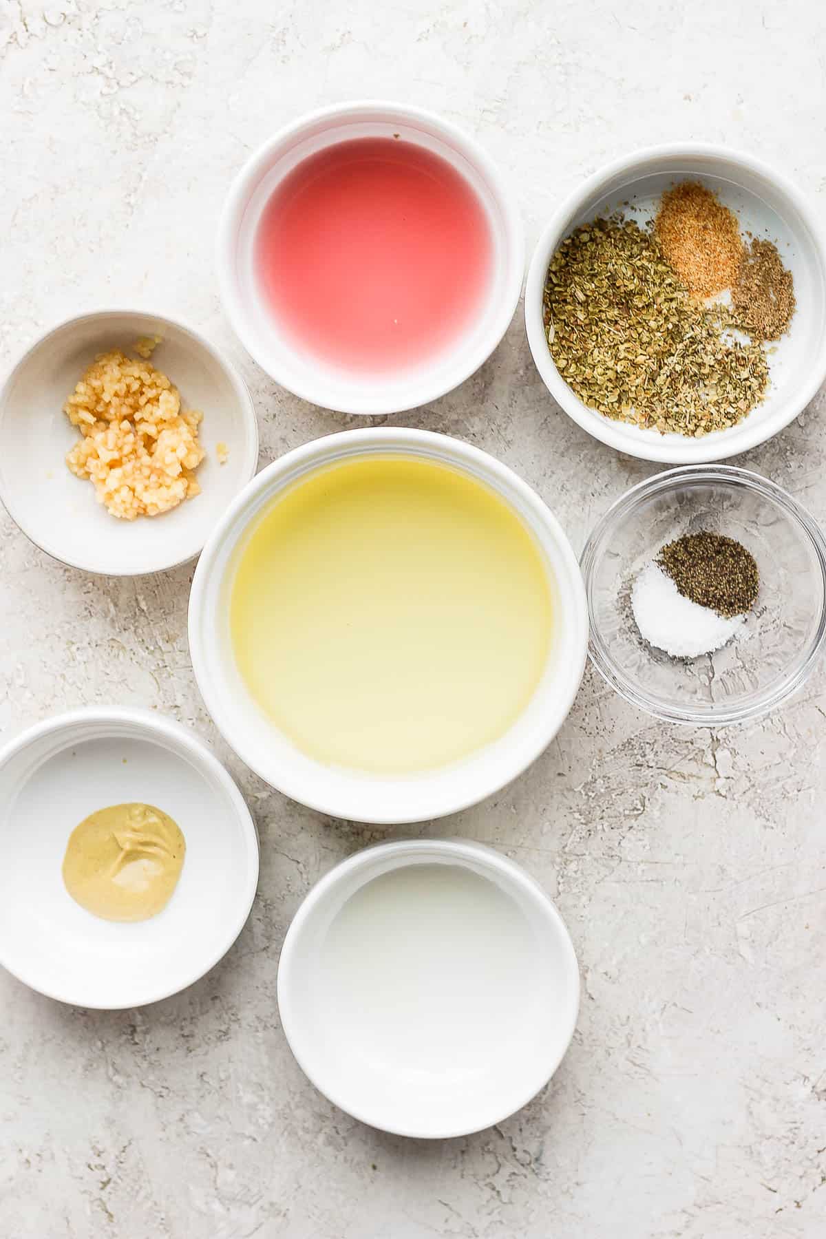 Seven small bowls, each with measured marinade ingredients; minced garlic, dijon mustard, salt and pepper, red wine vinegar, lemon juice, olive oil, and oregano, coriander, and garlic powder. 