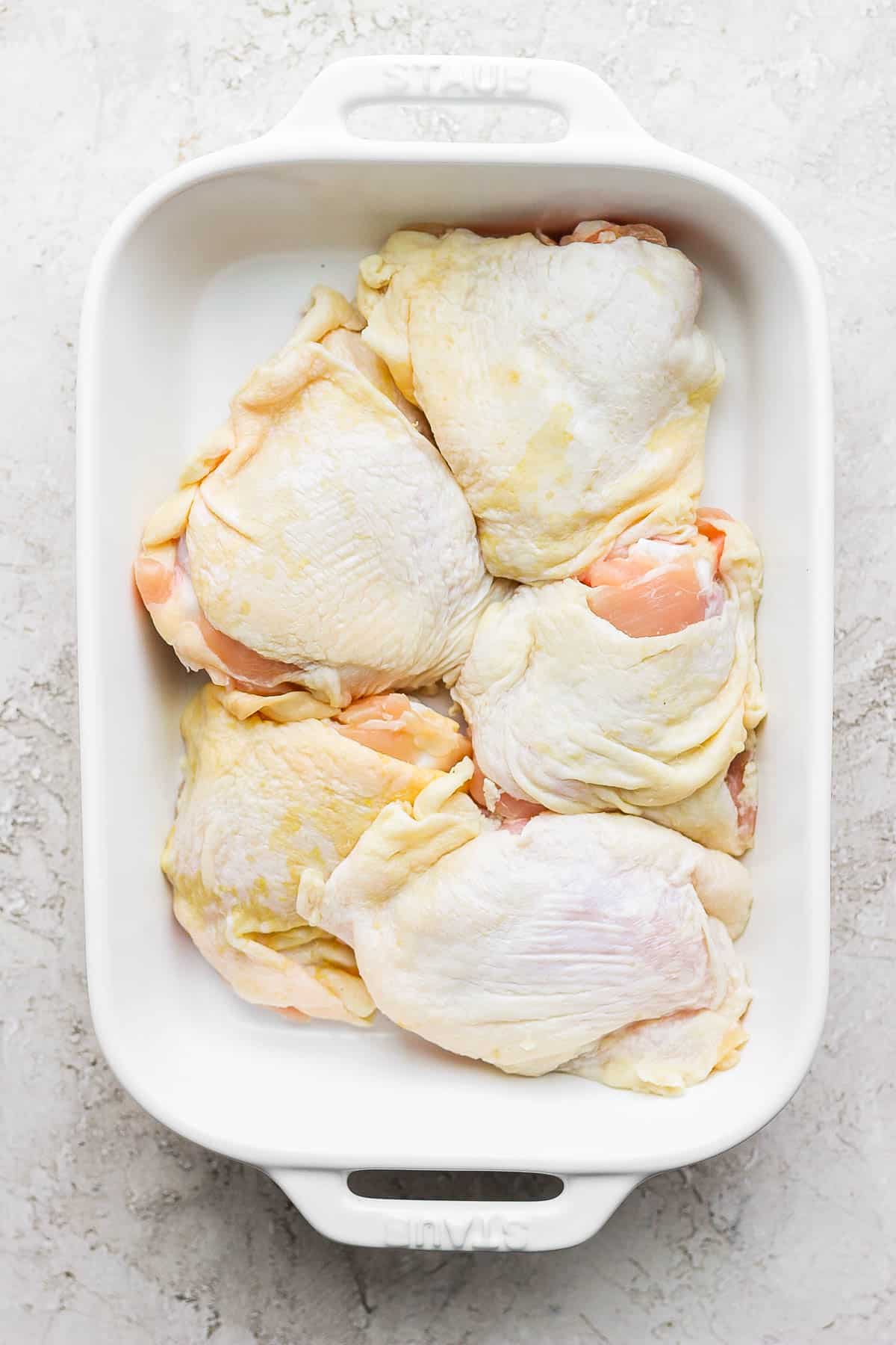 Five raw bone-in, skin on chicken thighs in a casserole dish.
