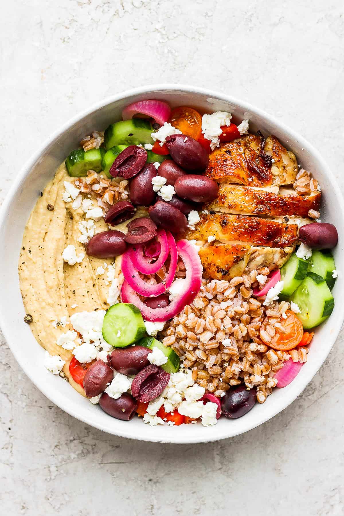 A bowl with a layer of hummus on the bottom topped with sliced English cucumbers, halved kalamata olives, pickled red onion, feta cheese crumbles, cooked farro, and a sliced greek chicken thigh. 