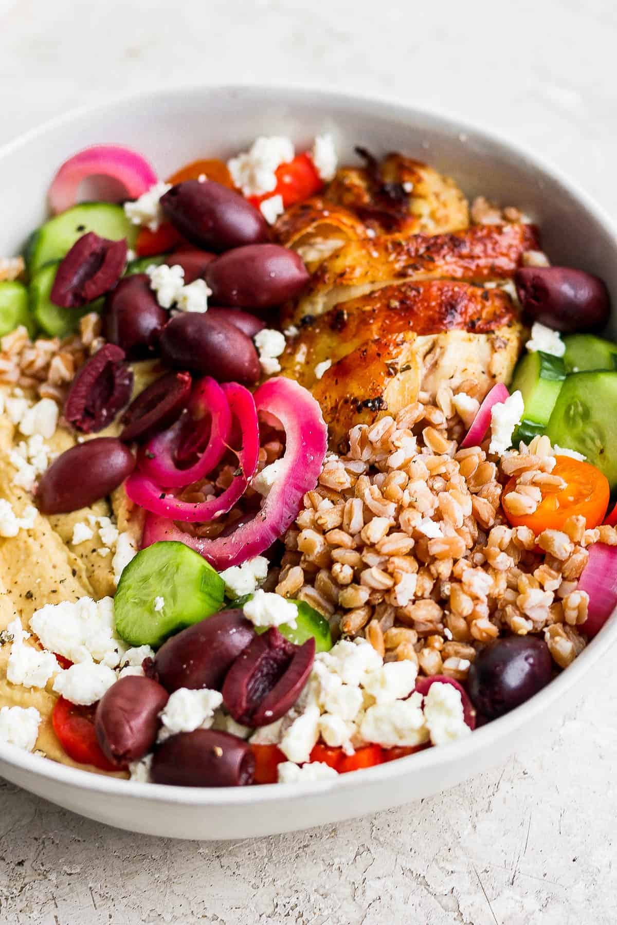 A bowl with a layer of hummus on the bottom topped with sliced English cucumbers, halved kalamata olives, pickled red onion, feta cheese crumbles, cooked farro, and a sliced greek chicken thigh. 