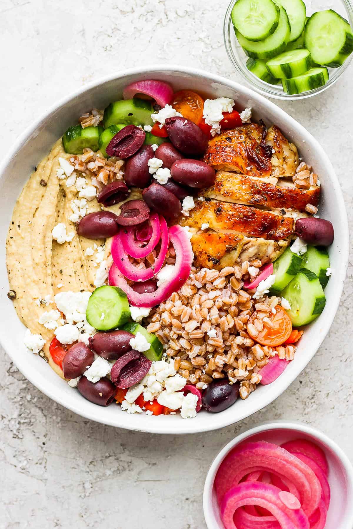 A bowl with a layer of hummus on the bottom topped with sliced English cucumbers, halved kalamata olives, pickled red onion, feta cheese crumbles, cooked farro, and a sliced greek chicken thigh. 