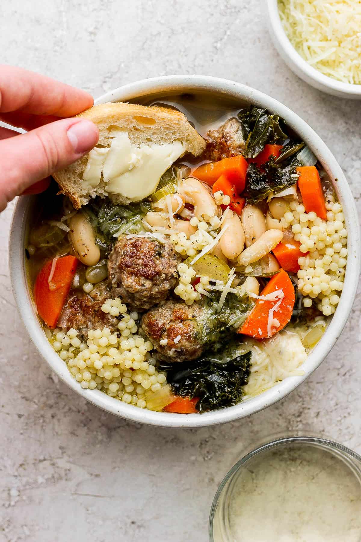 A hand dipping a slice of buttered sourdough bread in a bowl of Italian wedding soup.