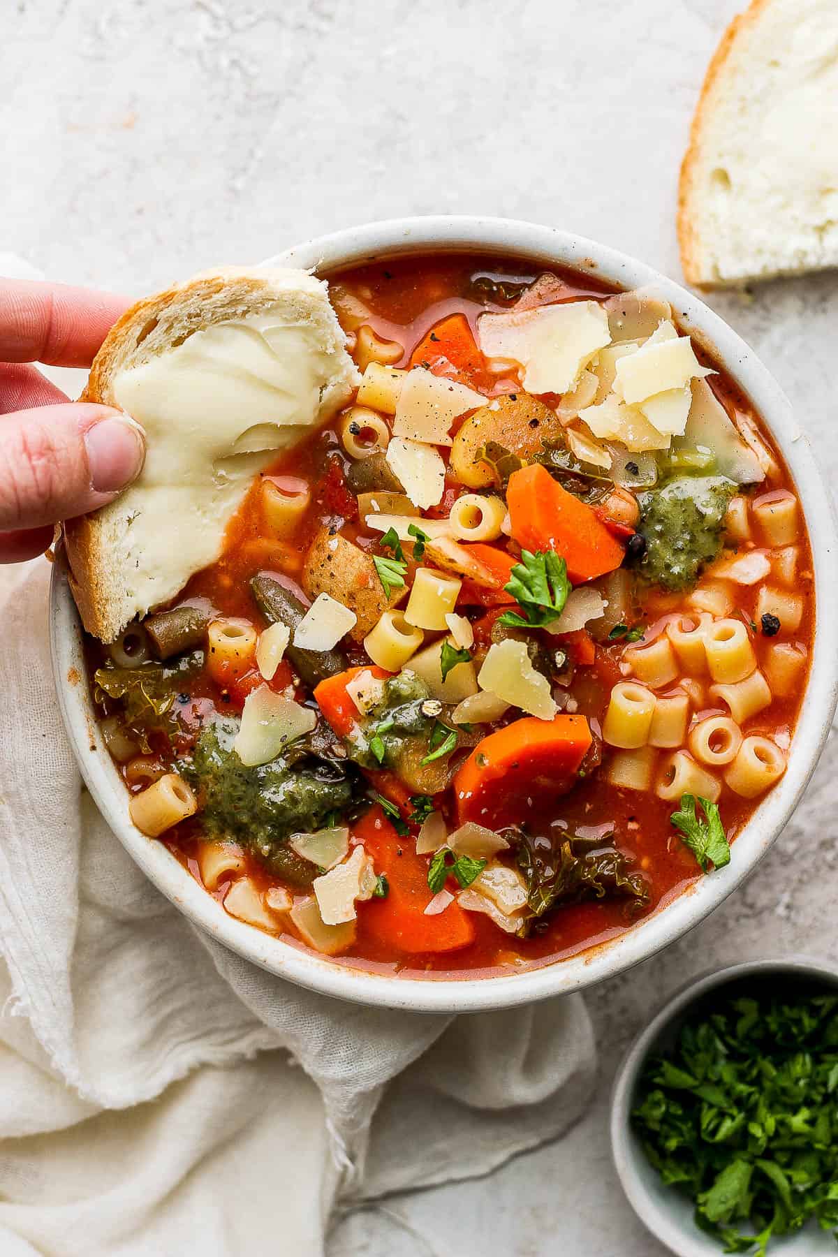 A slice of butter bread being dipped in a bowl of minestrone soup.