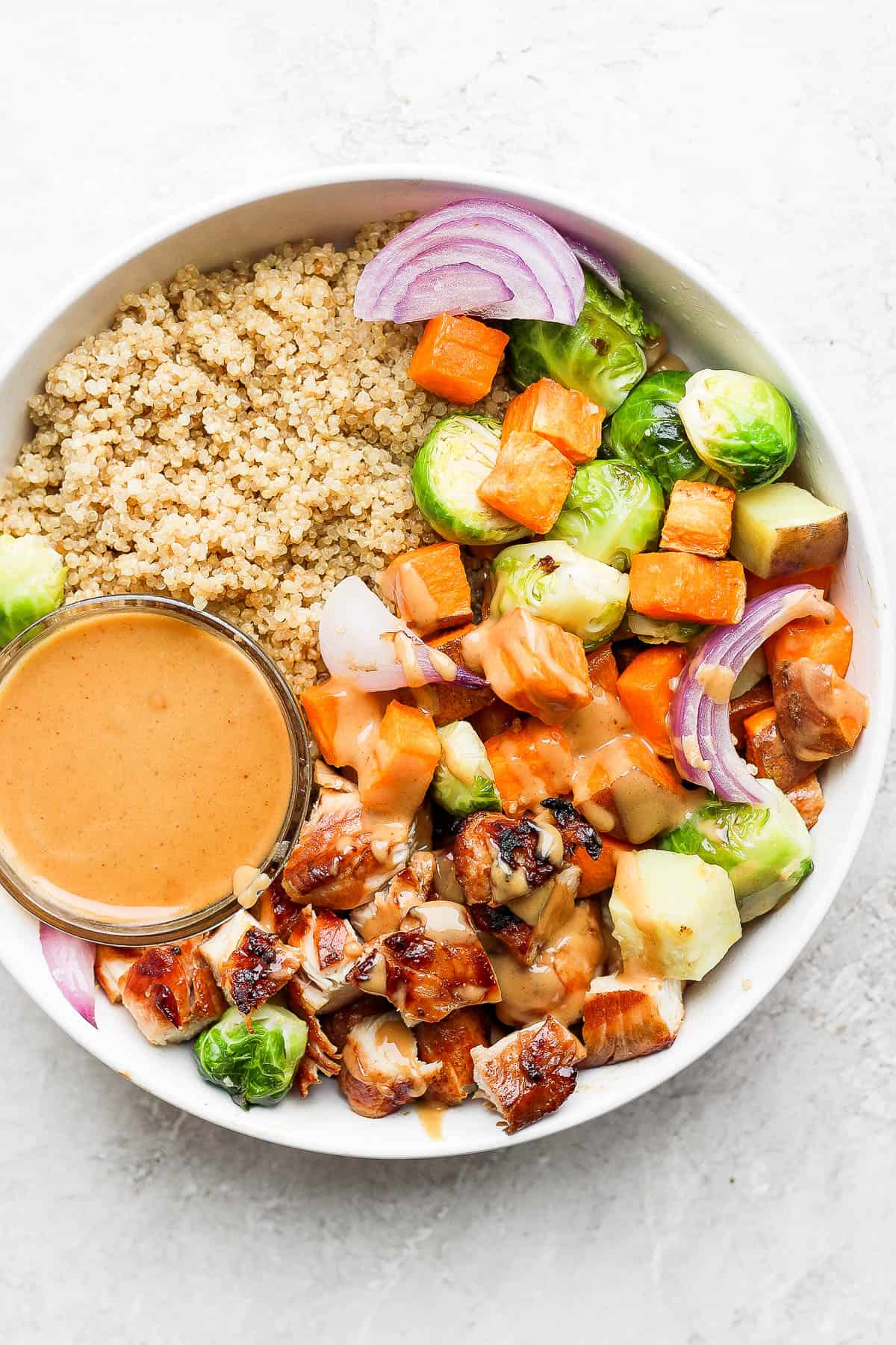A bowl with quinoa, roasted vegetables, grilled and cubed chicken, and a small bowl of peanut sauce. 