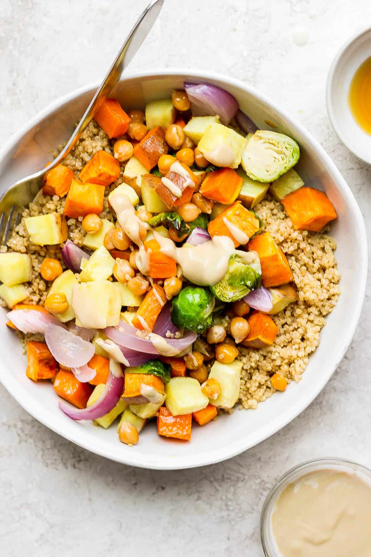 A bowl with a base of coconut quinoa topped with cooked brussels sprouts, cooked sweet potato, red onion, chickpeas, and a beige sauce. 