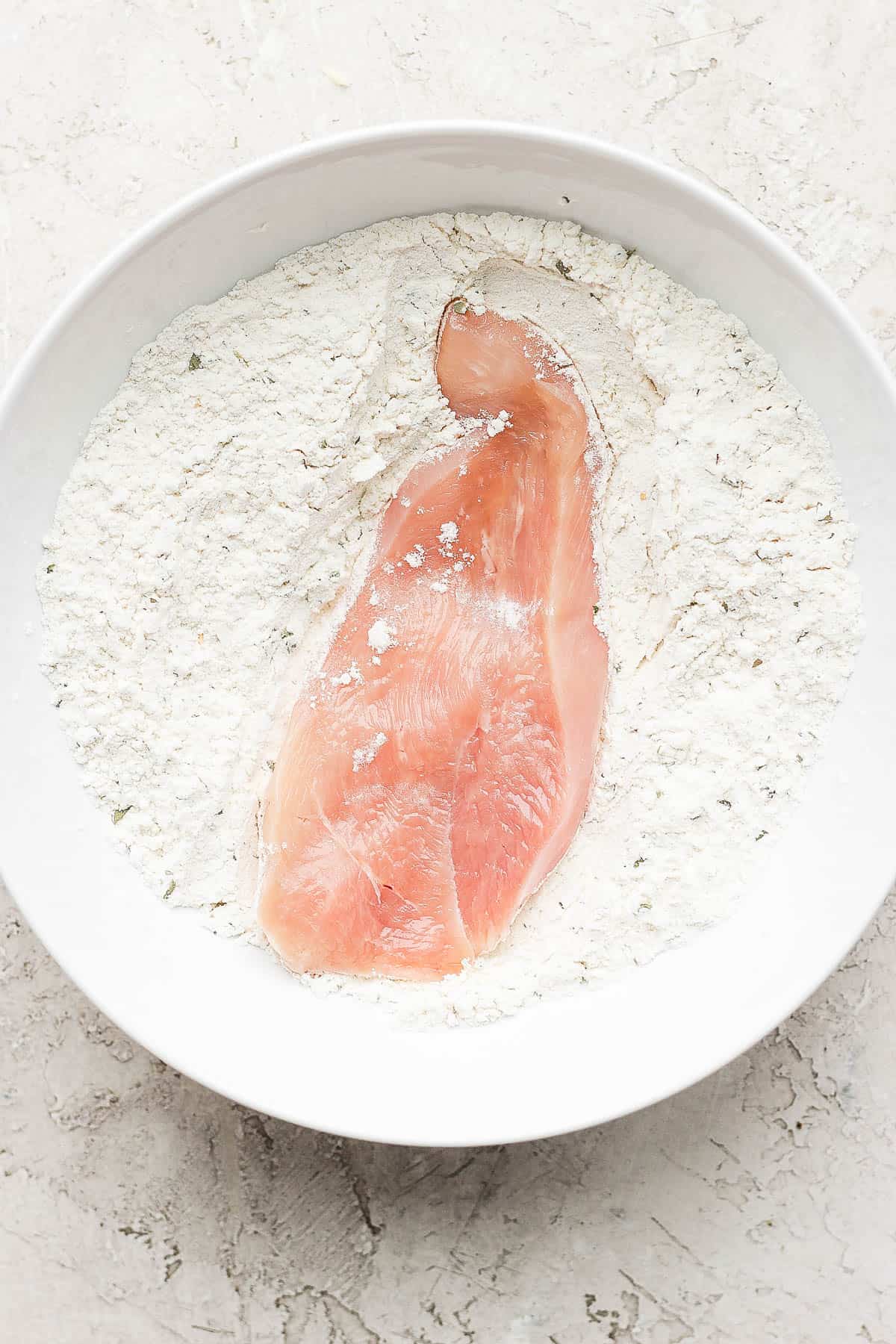 A chicken cutlet in the a white bowl full of flour, garlic powder, onion powder, and Italian seasoning.