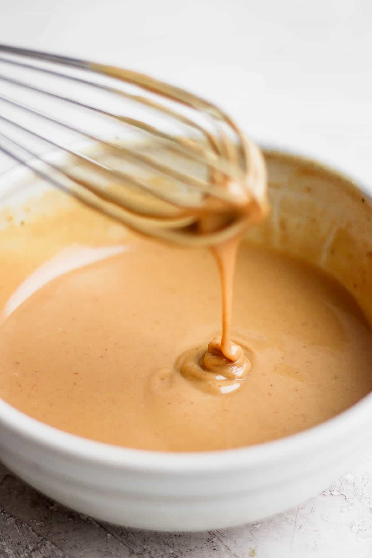 A whisk pulling out of the peanut sauce bowl with peanut sauce dripping off the end of the whisk to show a runny consistency. 