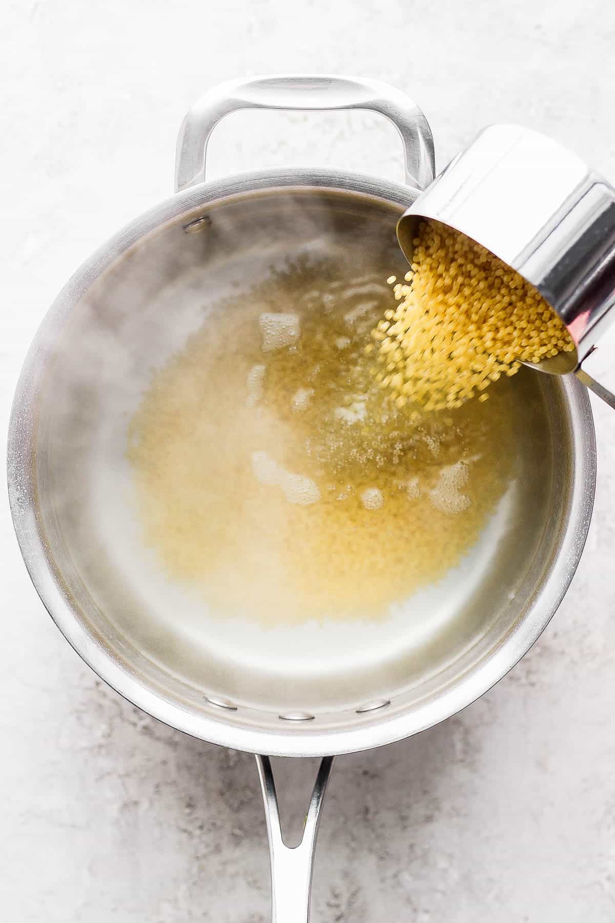 One cup of acini di pepe being poured from a measuring cup into a sauce pan full of steaming water.