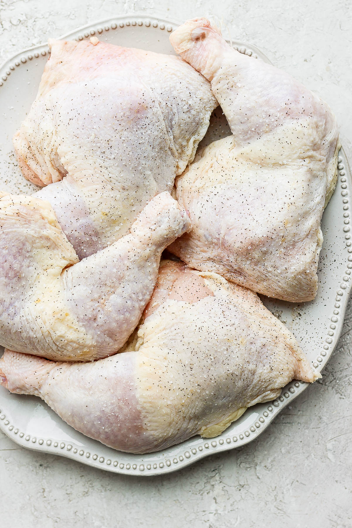 Raw chicken quarters on a plate that have been seasoned with salt and pepper.