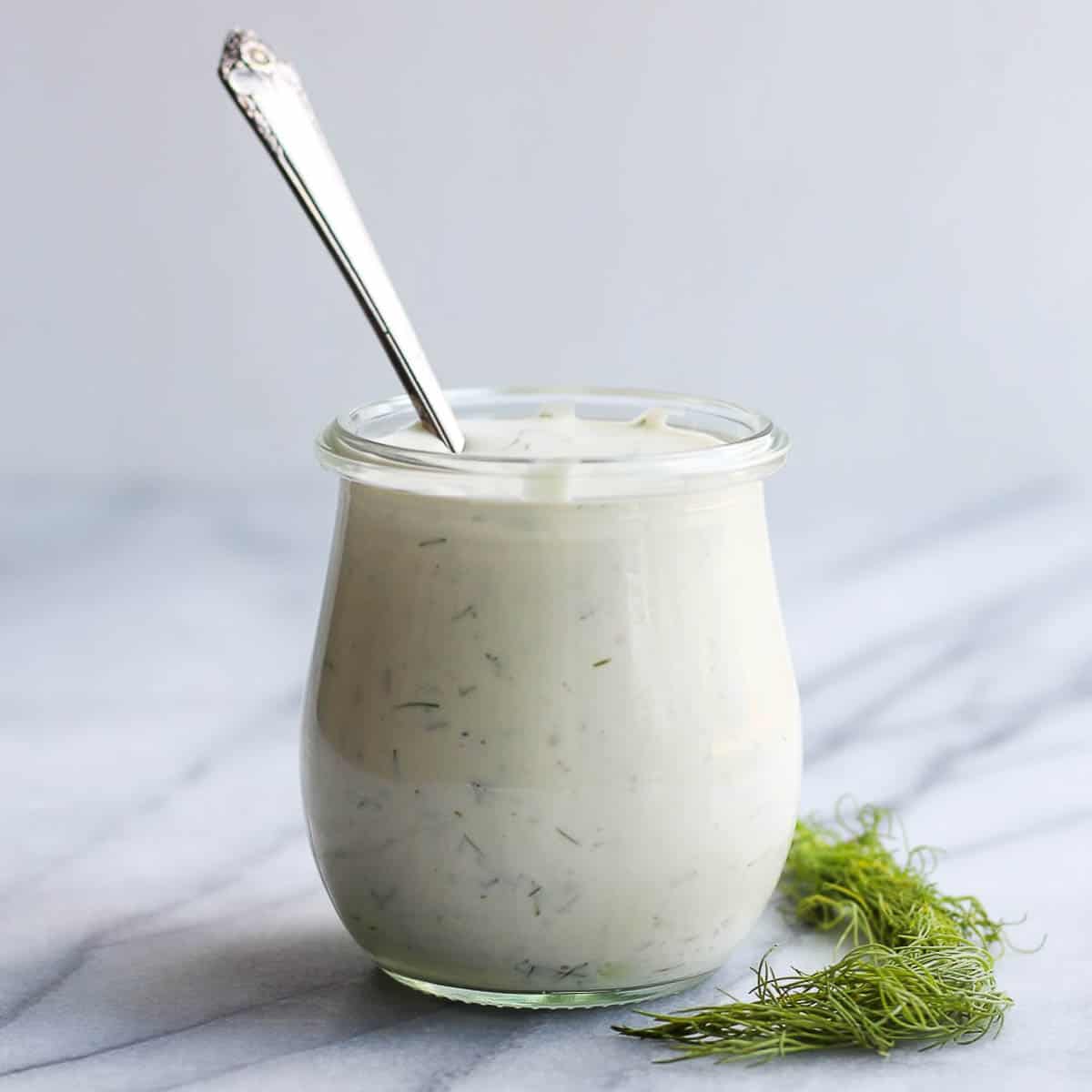 Tartar sauce in a glass jar with a spoon sticking out of the jar.  A handful of dill is sitting beside the jar.