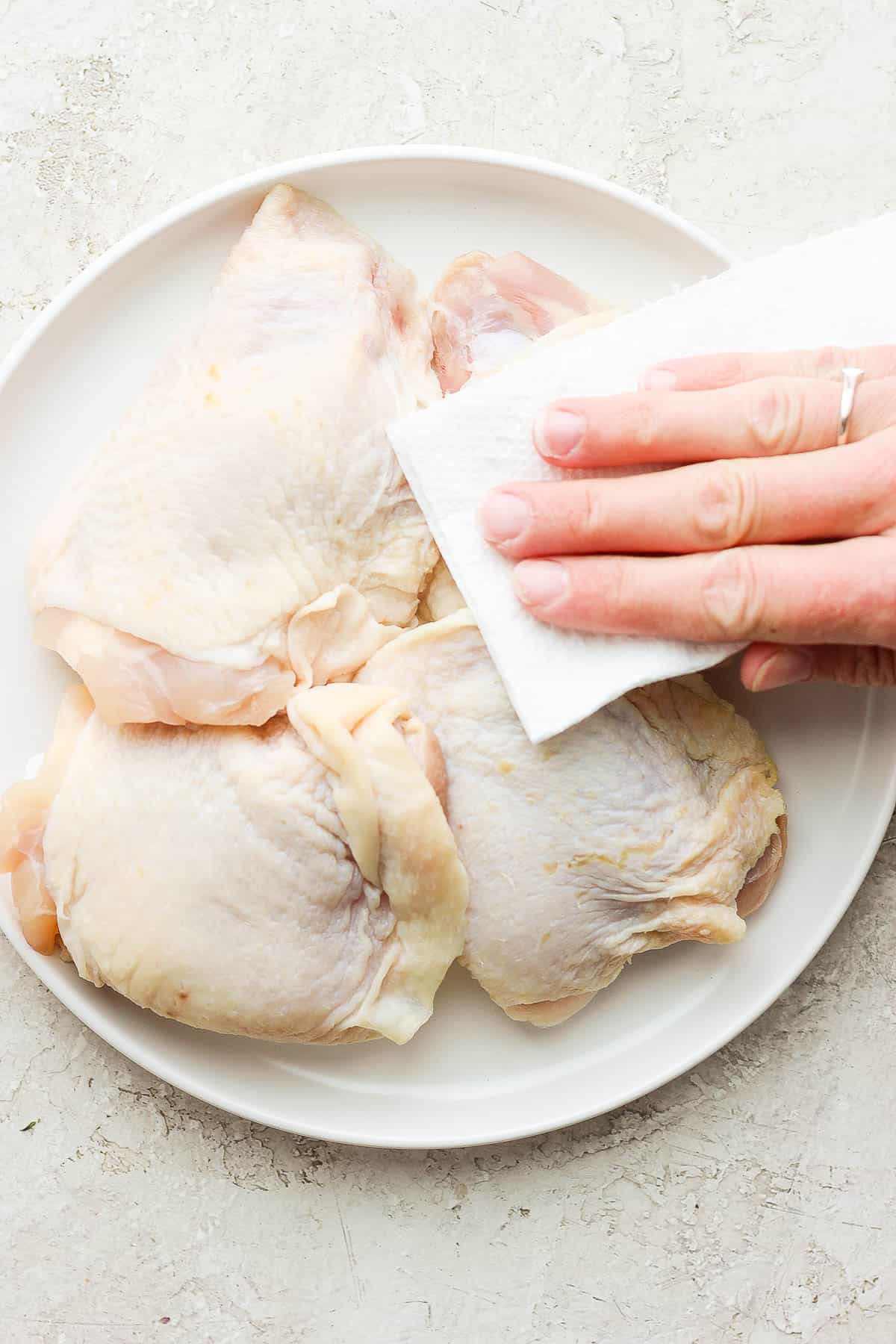 Raw chicken thighs on a white plate being pat dry with paper towel.