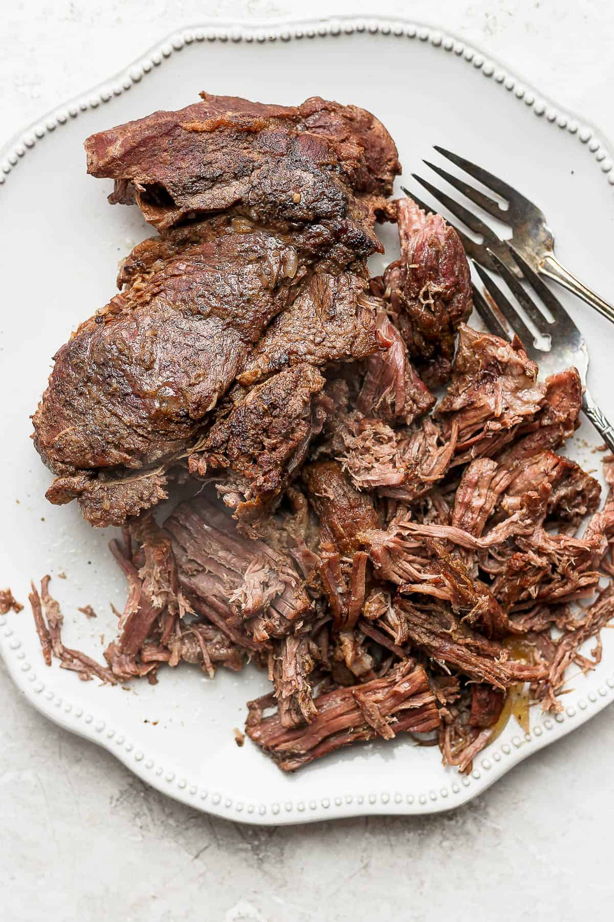 The cooked chuck roast on a plate and being shred by two forks.