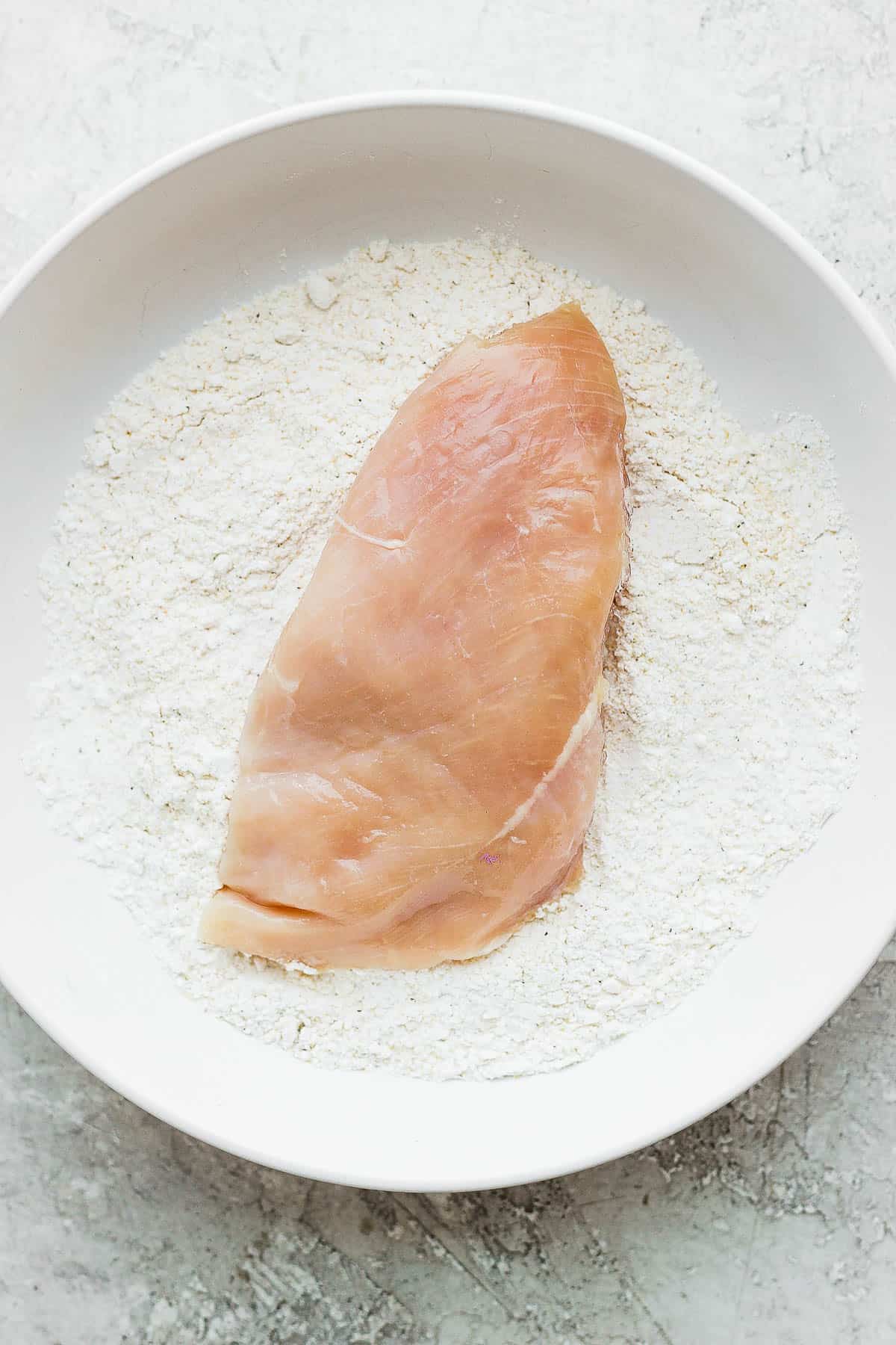 A raw chicken breast in a bowl with flour & seasonings.