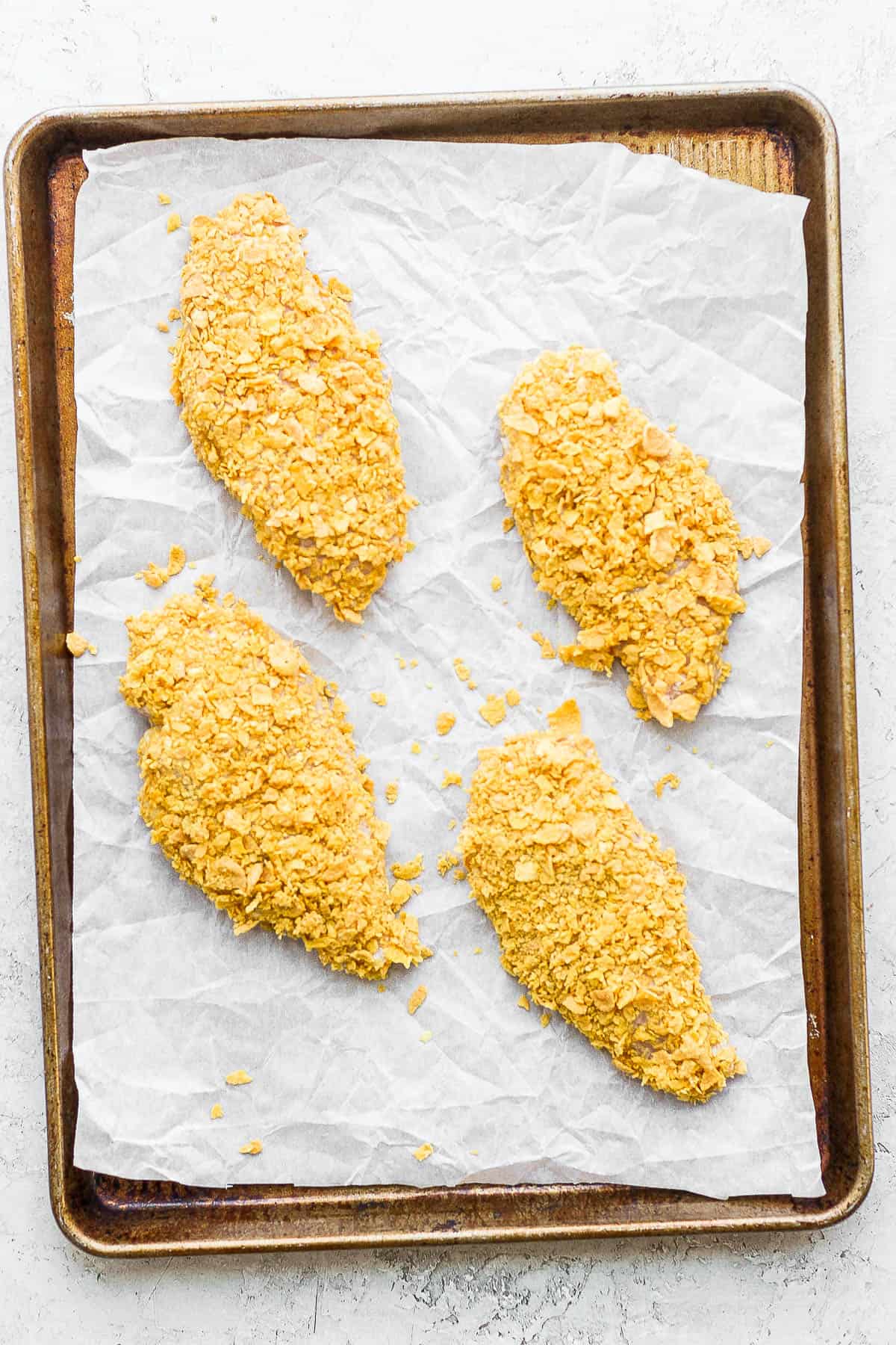 Cornflake-crusted chicken breasts on a parchment-lined baking sheet.