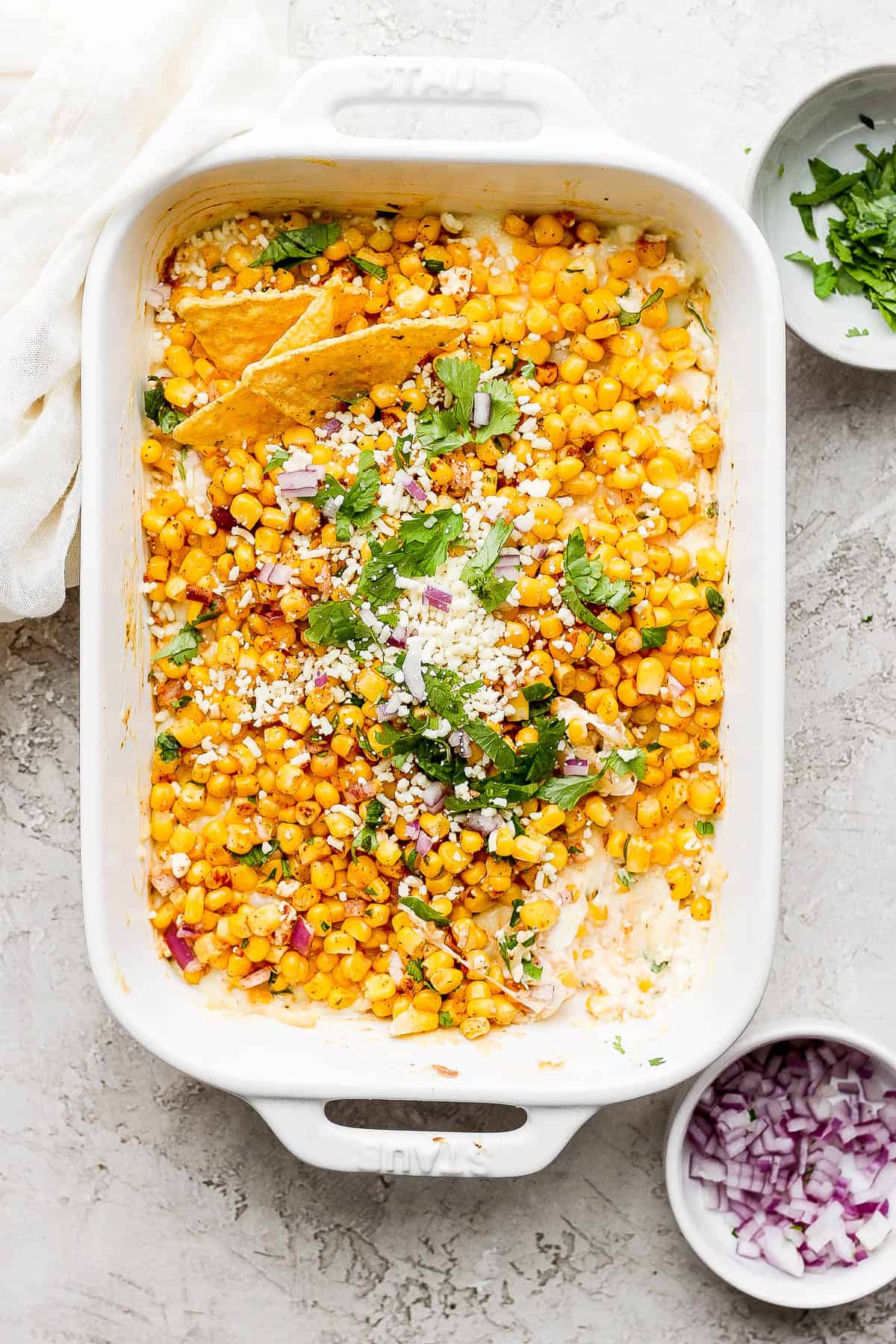 The same baking dish now topped with the remaining corn mixture, fresh cilantro, diced red onion, and crumbled cojita cheese.  Three chips are sticking into the dip.