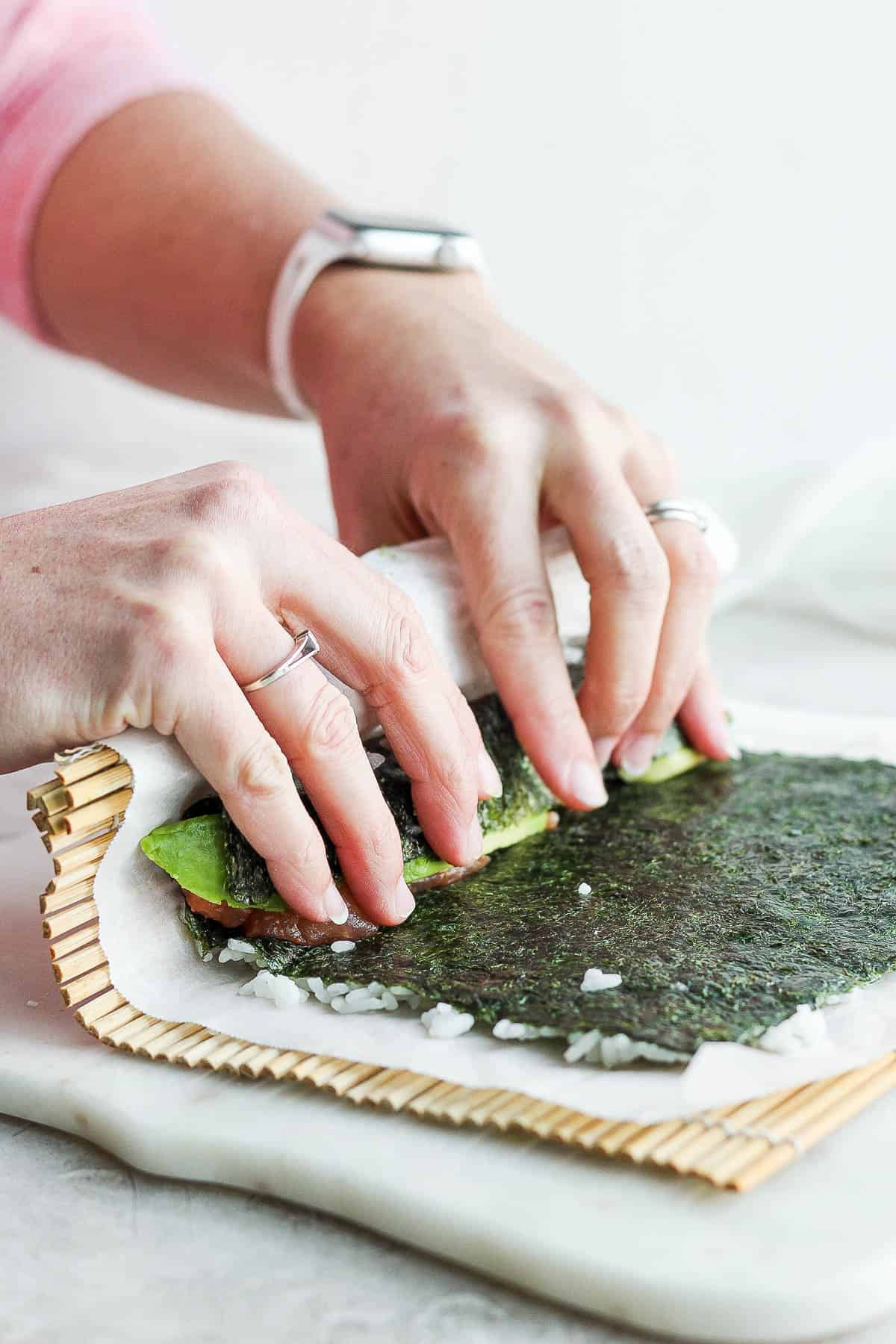 Two hands pulling the nori sheet over the filling ingredients.