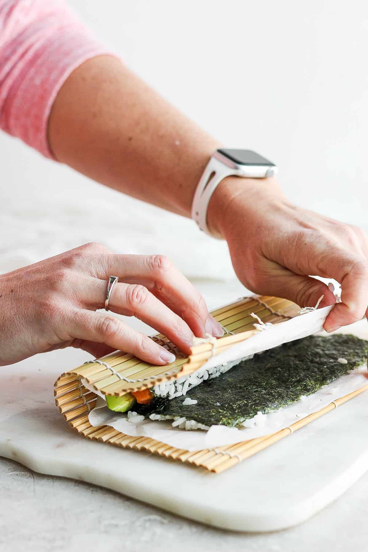 Two hands using the bamboo sheet to pull the nori sheet over the filling ingredients.