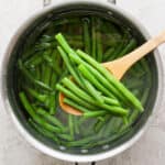 A saucepan of boiled green beans with a wooden spoon lifting a few out.